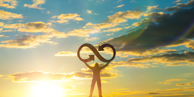 A silhouette of a man holds an infinity symbol on top of a mountain with a sunset background. Concept idea, life, success, business.