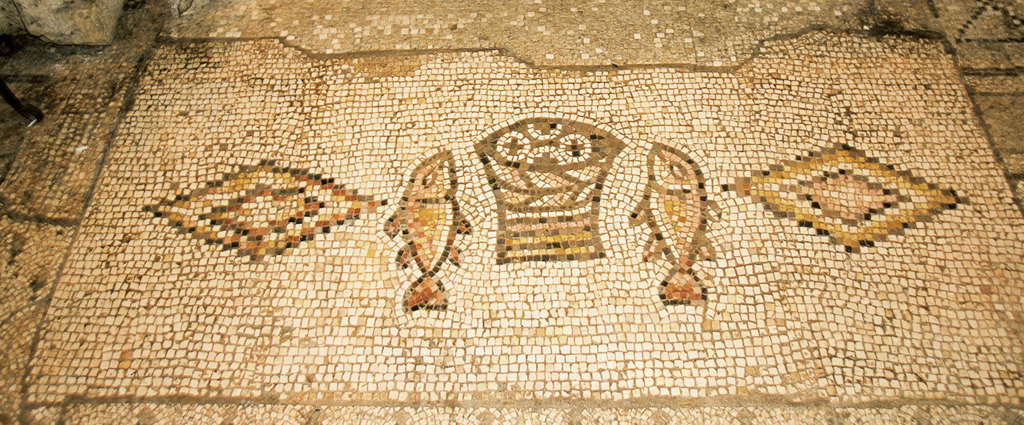 Das berühmte Brot- und Fisch-Mosaik in der Brotvermehrungskirche Tabgha am See Genezareth in Israel: Aus einem Korb ragen vier runde, mit Kreuzen gezeichneten Brote hervor. Links und rechts davon liegt ein Fisch, der gedrungenen Form nach wohl ein Pe