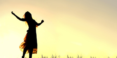 Frau steht im Gegenlicht vor einem gelben Himmel und reckt die Arme jubelnd nach oben